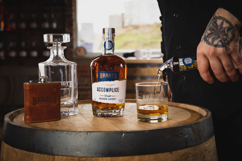 Brady's Accomplice Bourbon, flask, and decanter on a barrel while whiskey is poured